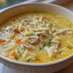 Close-up of a bowl filled with rich, yellow Creamy Lemon Chicken Orzo Soup, featuring shredded chicken, orzo pasta, and dill.