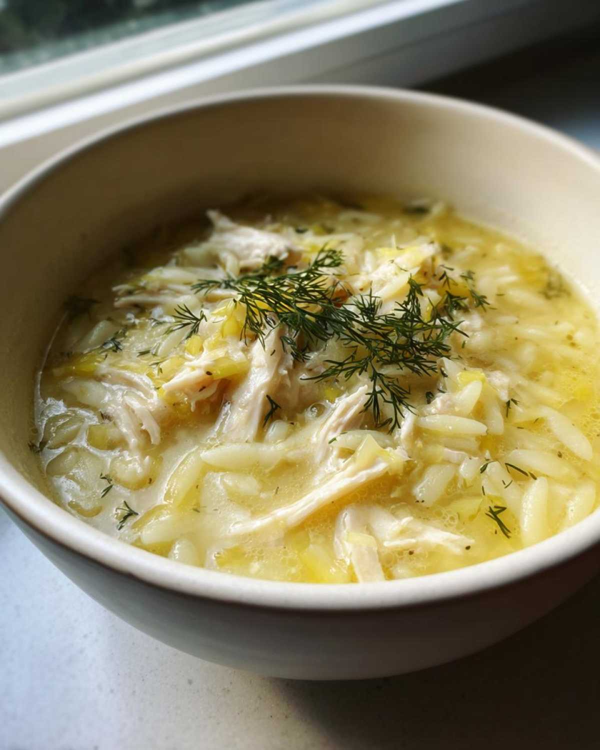 Close-up of a bowl of Creamy Lemon Chicken Orzo Soup, topped with shredded chicken and fresh dill.