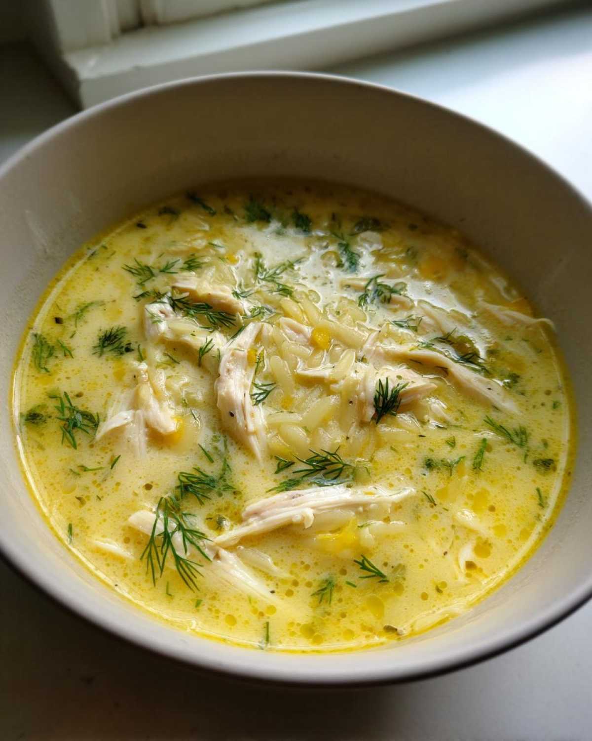 Close-up of a bowl of rich, yellow Creamy Lemon Chicken Orzo Soup garnished with fresh dill.
