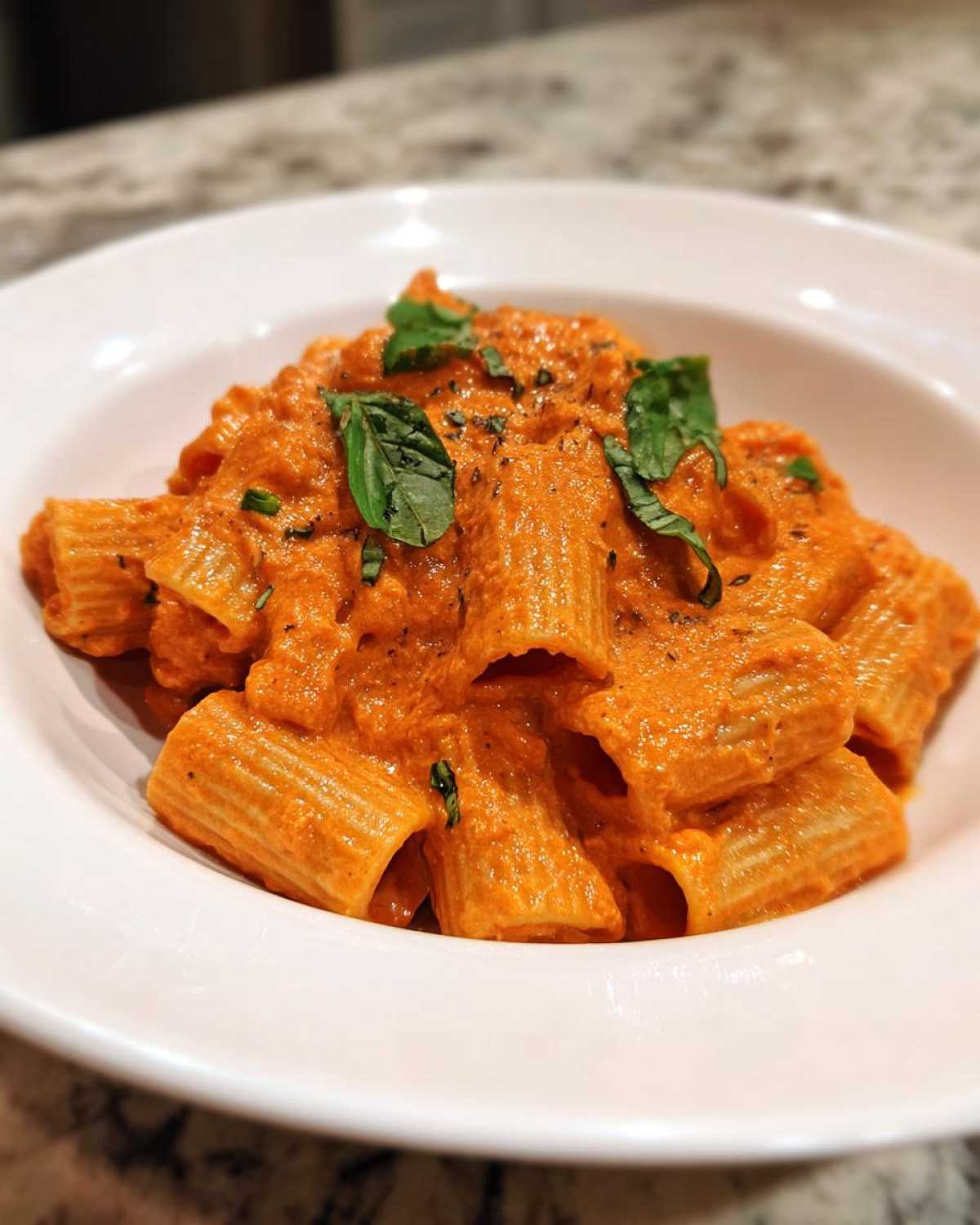 Close-up of rigatoni pasta coated in vibrant orange Creamy Roasted Red Pepper Pasta sauce, garnished with fresh basil.