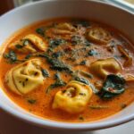 Close-up of a white bowl filled with rich, orange Creamy Tomato Tortellini Soup featuring plump tortellini and wilted spinach.