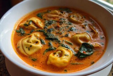 Close-up of a white bowl filled with rich, orange Creamy Tomato Tortellini Soup featuring plump tortellini and wilted spinach.