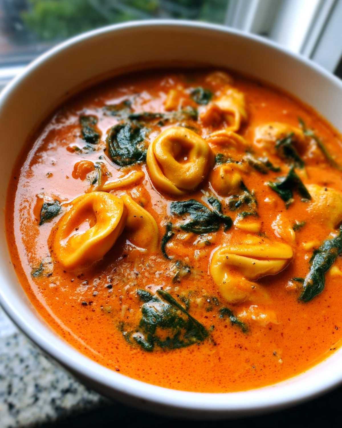 Close-up of a white bowl filled with rich, orange Creamy Tomato Tortellini Soup featuring cheese tortellini and wilted spinach.