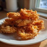 A close-up of a pile of golden brown, super crispy Air Fryer Onion Rings stacked on a white plate.
