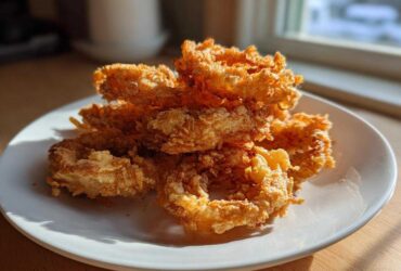 A close-up of a pile of golden brown, super crispy Air Fryer Onion Rings stacked on a white plate.