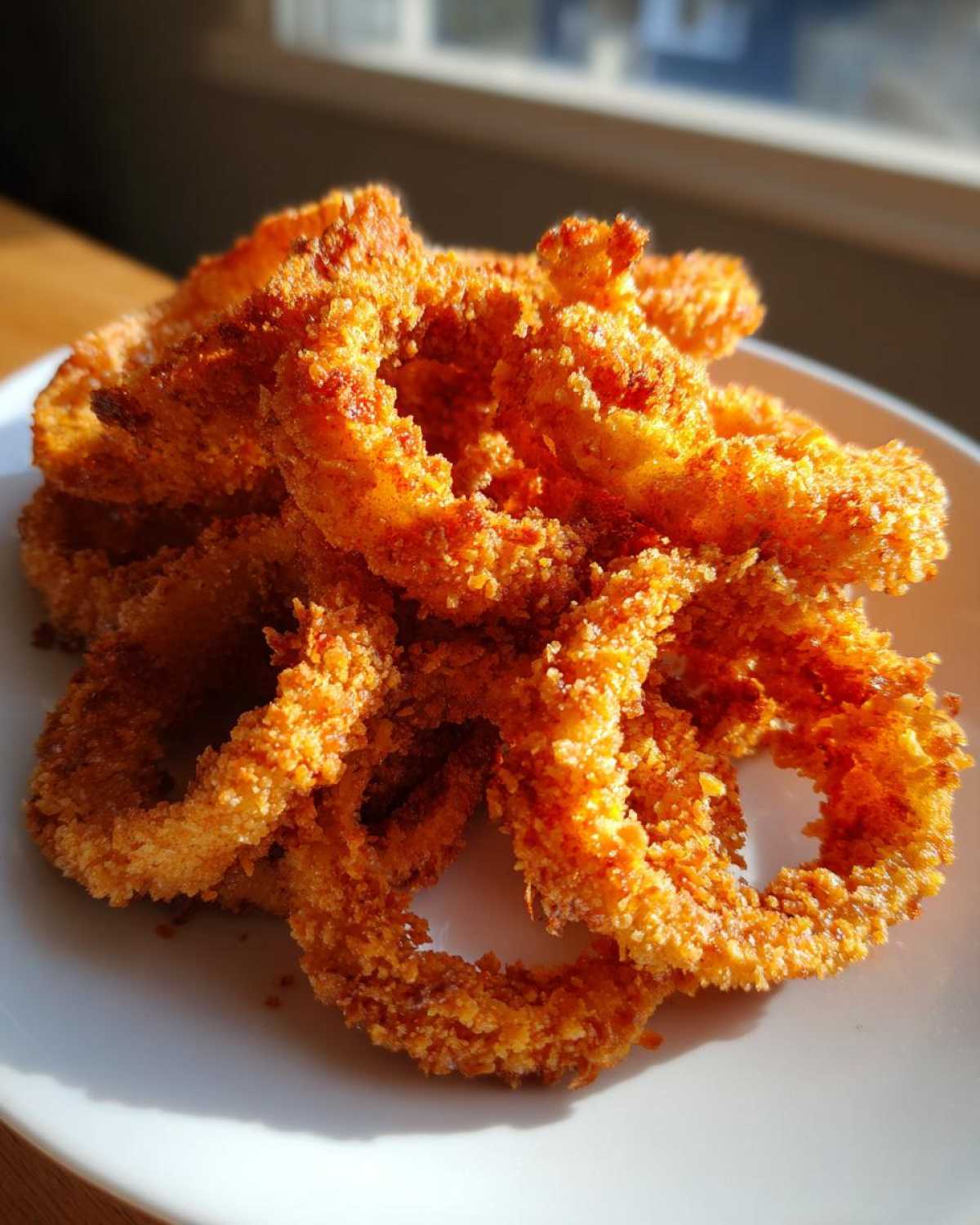 A close-up pile of golden brown, crispy Air Fryer Onion Rings catching bright sunlight on a white plate.