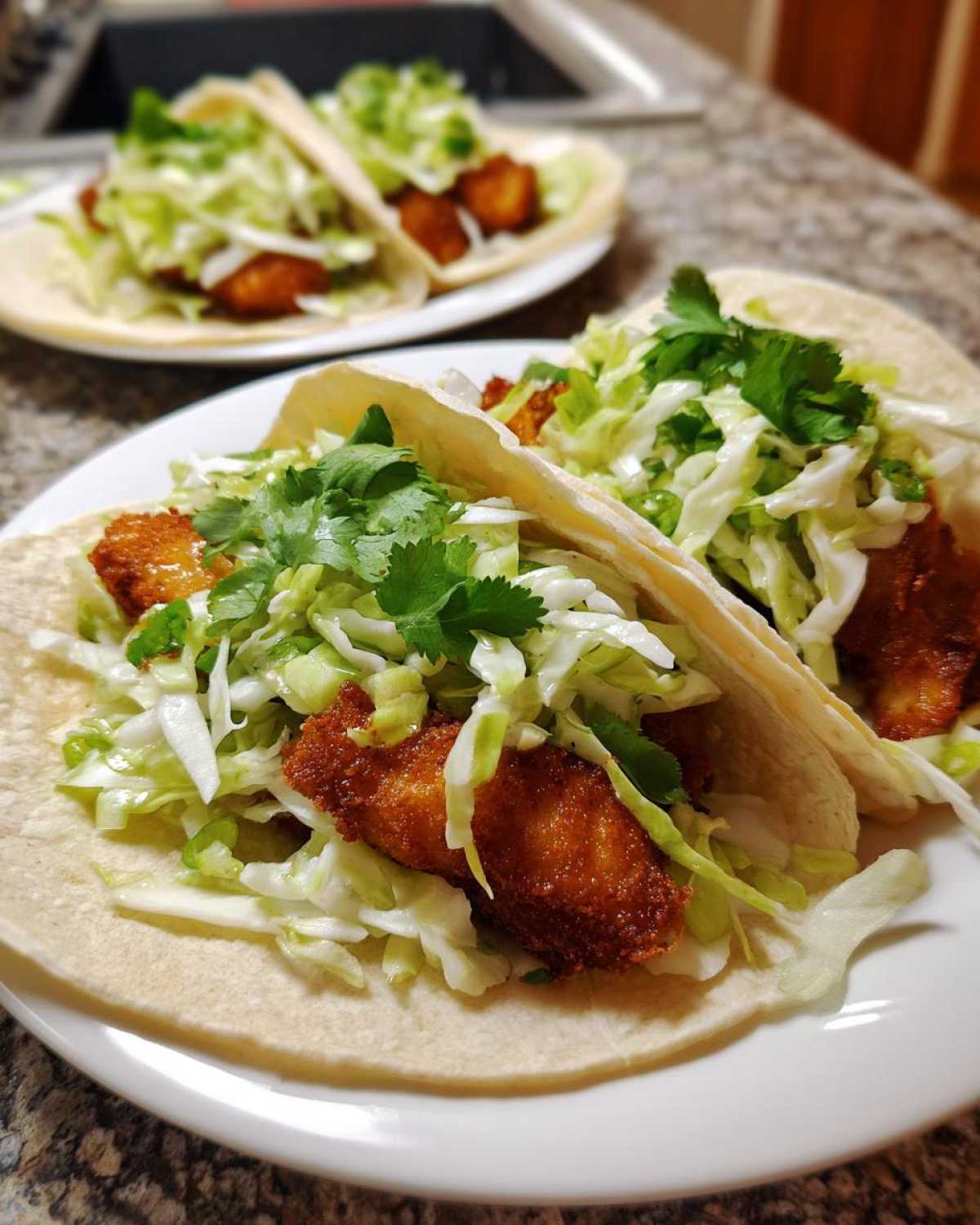 Two Crispy Baked Fish Tacos filled with golden fried fish pieces, shredded lime slaw, and cilantro on a white plate.