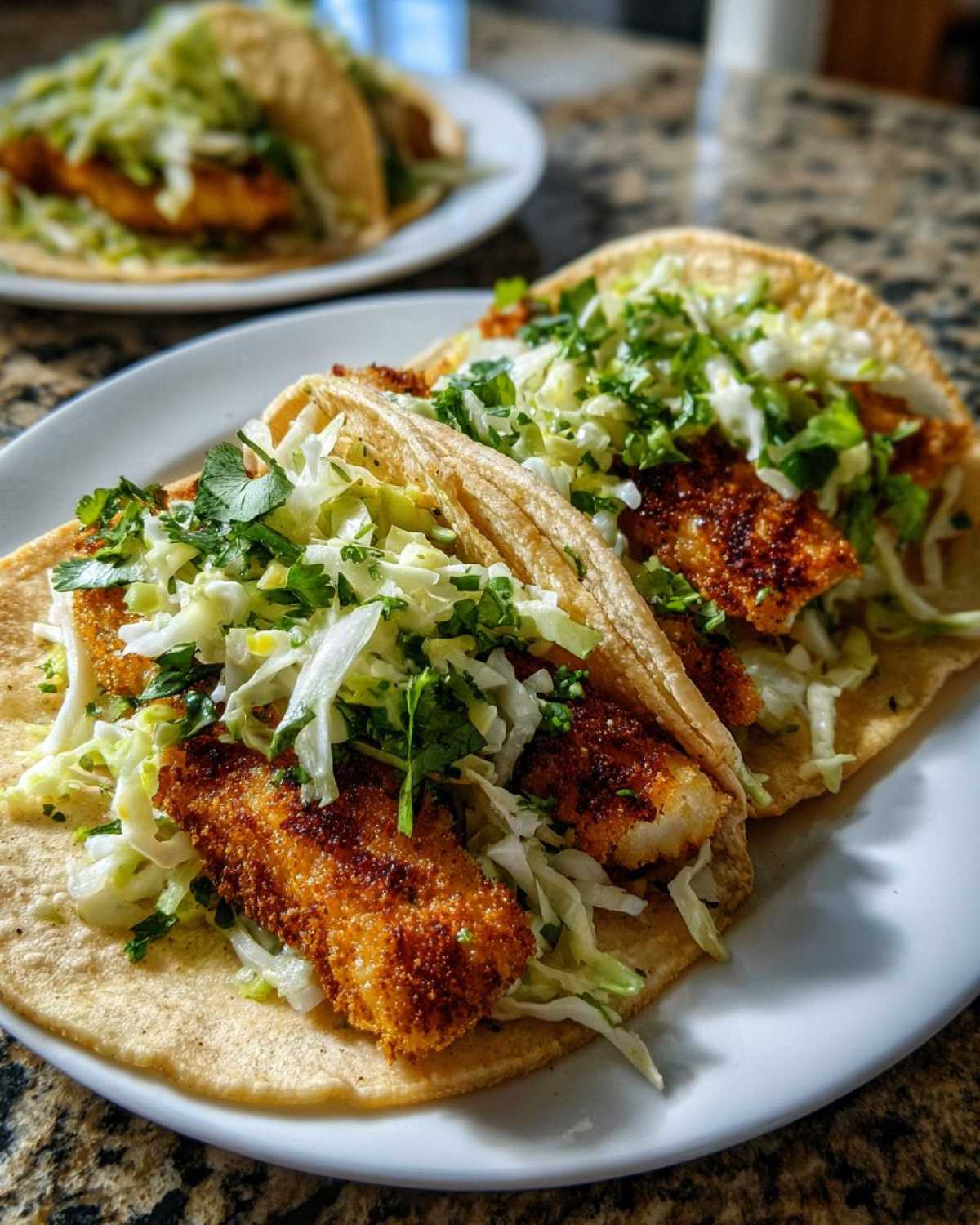 Two Crispy Baked Fish Tacos topped with shredded cabbage slaw and cilantro on a white plate.