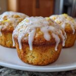 Three freshly baked Glazed Lemon Poppy Seed Muffins with thick white icing dripping down the sides, served on a white plate.