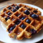 Close-up of two golden brown Gluten-Free Blueberry Waffles topped with fresh blueberries and syrup.