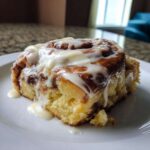 A single, fluffy Gluten-Free Cinnamon Roll generously topped with white cream cheese icing, sitting on a white plate.