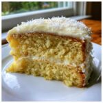 Close-up of a moist slice of Gluten-Free Lemon Coconut Cake layered with cream and topped with white frosting and shredded coconut.