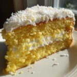 Close-up of a moist slice of Gluten-Free Lemon Coconut Cake layered with cream and topped with shredded coconut.
