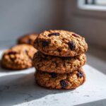 A stack of three chewy Gluten-Free Oatmeal Raisin Cookies featuring visible raisins, set against a bright, sunlit background.