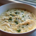 Close-up of a bowl of creamy Greek Lemon Chicken Soup featuring shredded chicken and orzo pasta, garnished with parsley.