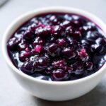 A close-up of thick, glossy Homemade Blueberry Syrup filling a small white bowl, featuring plump, cooked blueberries.