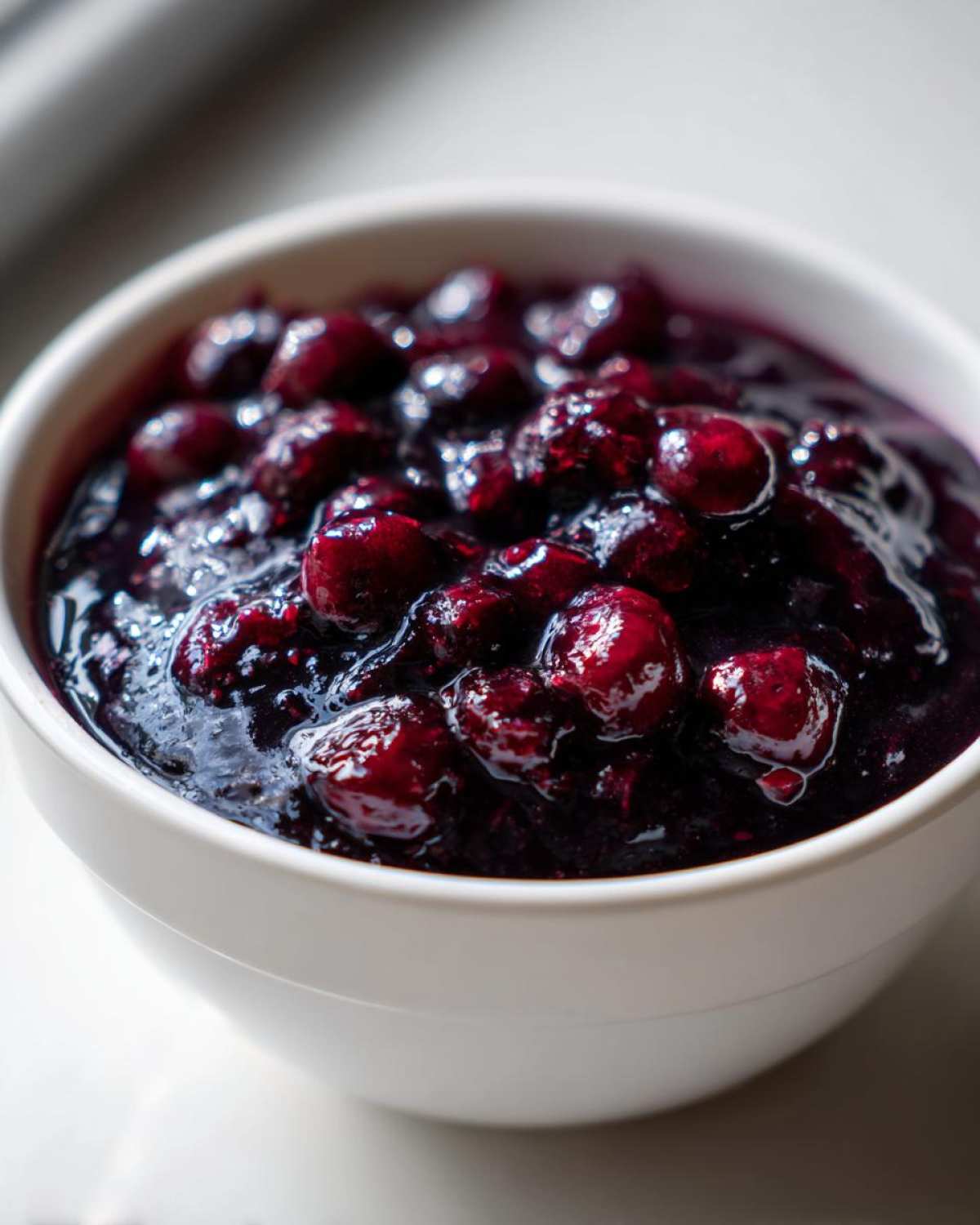 A close-up of thick, rich Homemade Blueberry Syrup with plump, glossy whole blueberries in a white bowl.