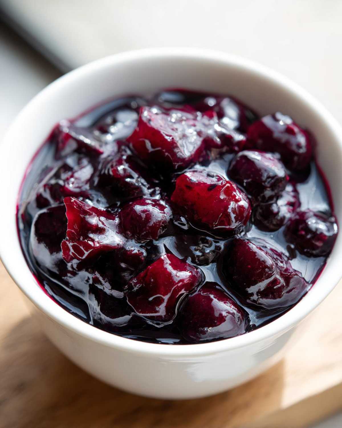 Close-up of thick, dark purple homemade blueberry syrup filled with plump, glistening whole blueberries in a white bowl.