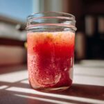 A glass jar filled with vibrant red Homemade Strawberry Lemonade Concentrate, showing texture and lemon zest on top.