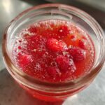 Close-up overhead view of bright red Homemade Strawberry Lemonade Concentrate with whole strawberries visible inside a glass jar.