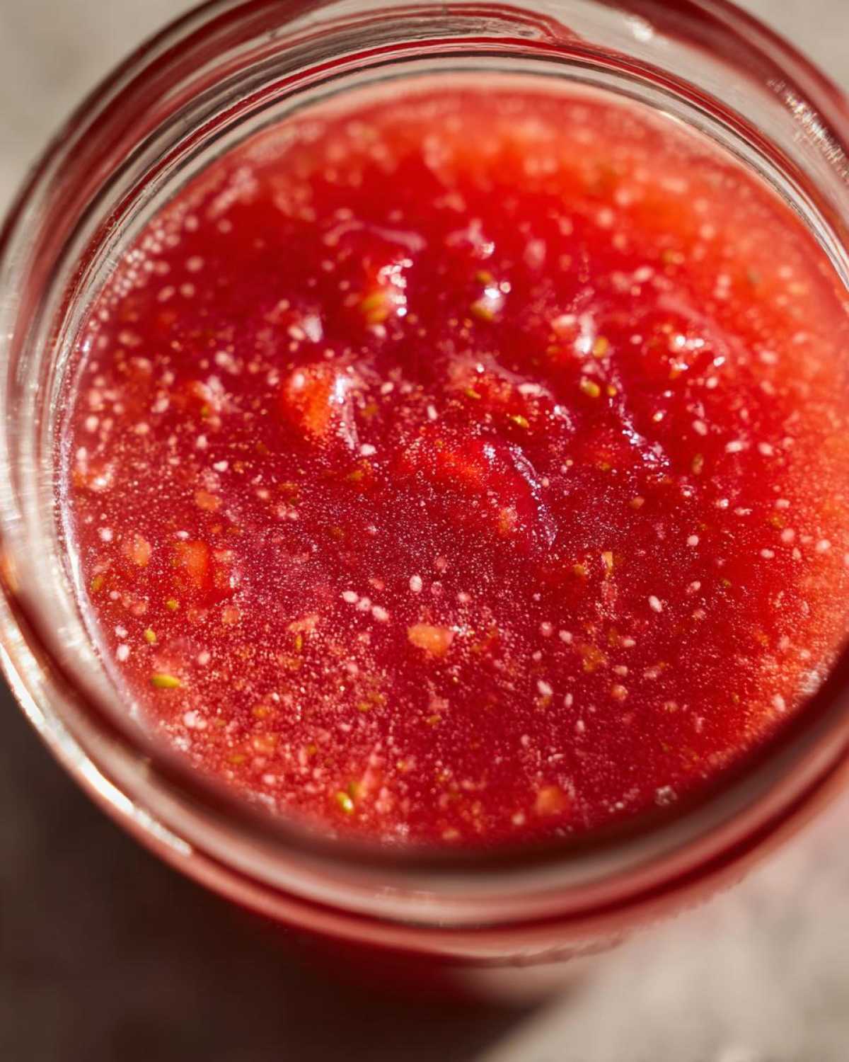 Top-down view of vibrant red Homemade Strawberry Lemonade Concentrate in a glass jar, showing pulp and seeds.