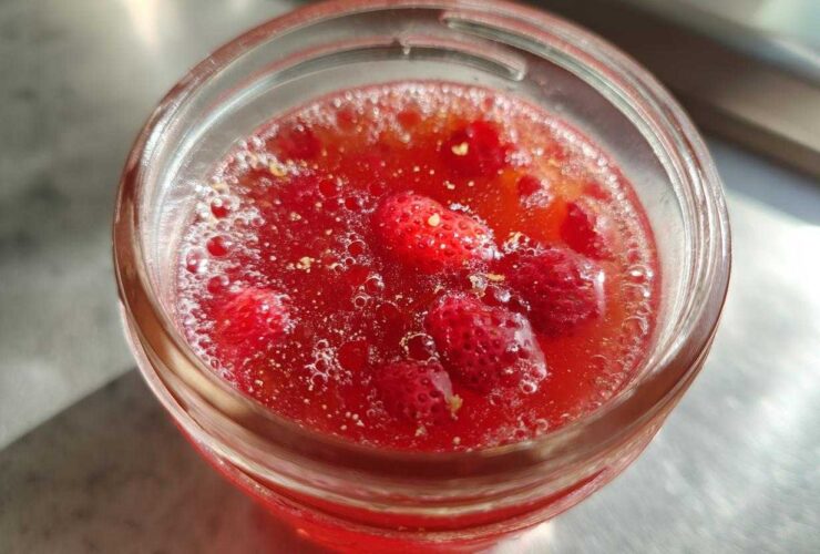 Close-up overhead view of bright red Homemade Strawberry Lemonade Concentrate with whole strawberries visible inside a glass jar.