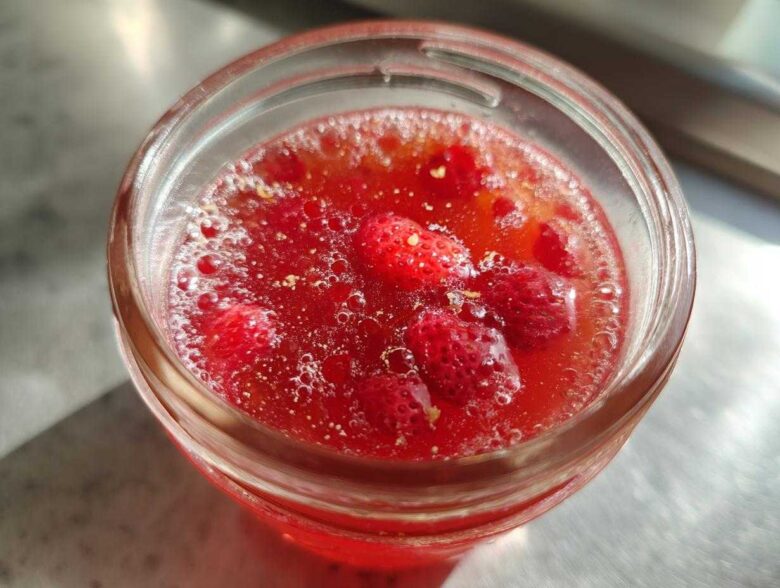 Close-up overhead view of bright red Homemade Strawberry Lemonade Concentrate with whole strawberries visible inside a glass jar.