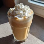 A tall glass filled with a creamy Homemade Vanilla Bean Frappuccino, topped high with whipped cream.