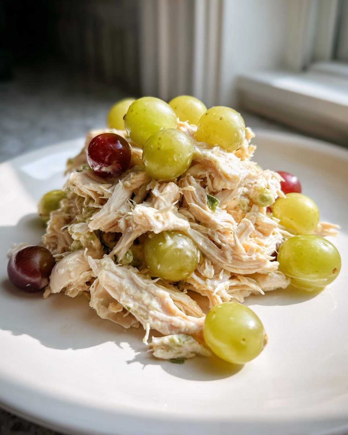 A mound of shredded Honey Mustard Chicken Salad with Grapes on a white plate, brightly lit.