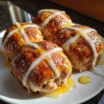 Four freshly baked Hot Cross Buns with Orange Glaze, drizzled with icing and syrup, sitting on a white plate.