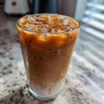 A tall, sweating glass filled with a layered Iced Brown Sugar Oatmilk Latte, topped with ice cubes.