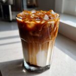 Close-up of a tall glass filled with a layered Iced Caramel Macchiato, topped with ice and rich caramel drizzle.