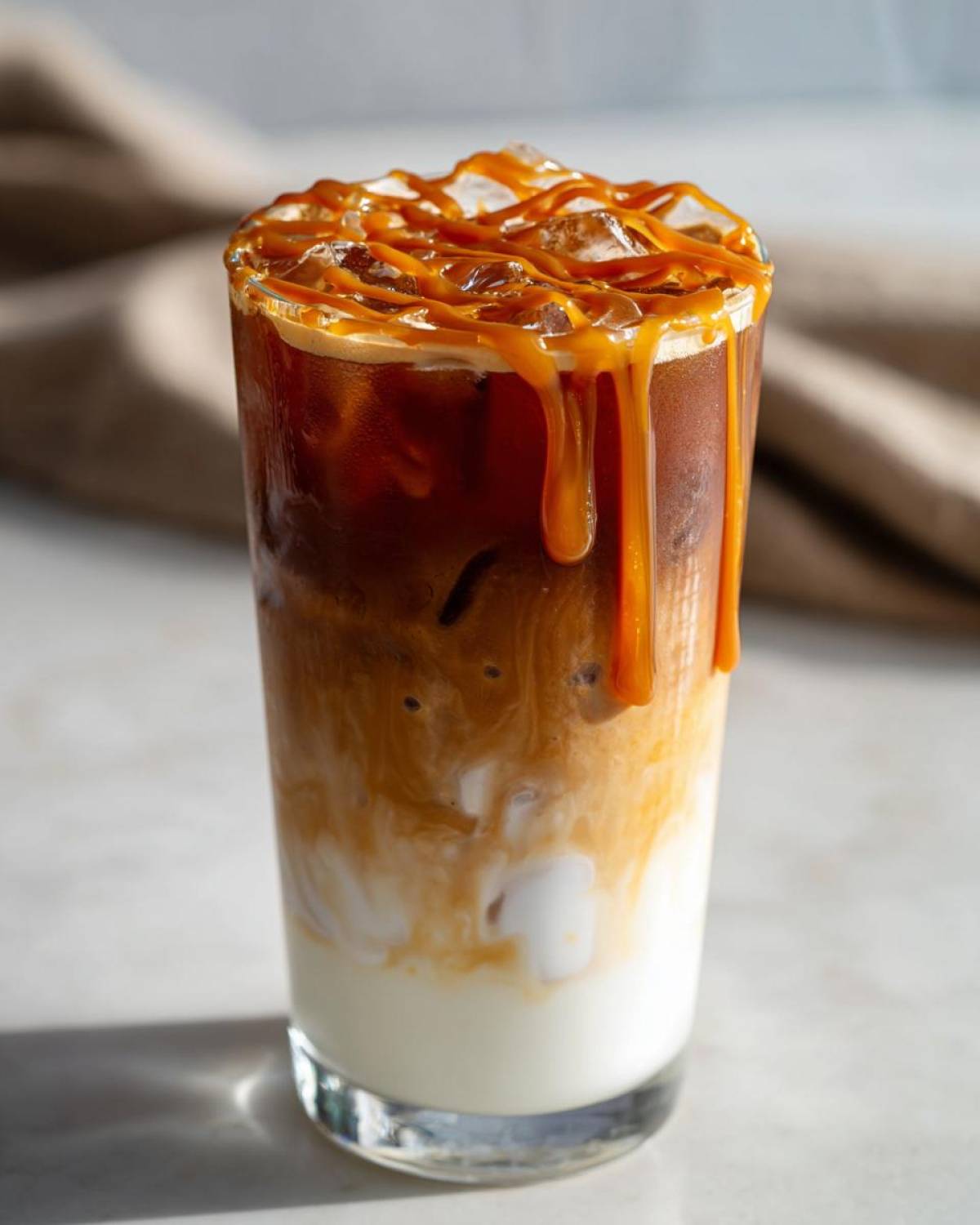 Close-up of a tall glass featuring a layered Iced Caramel Macchiato with milk, espresso, and heavy caramel drizzle over ice.