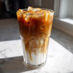 Tall glass filled with an Iced Caramel Macchiato showing distinct layers of milk, espresso, and caramel drizzle over ice.