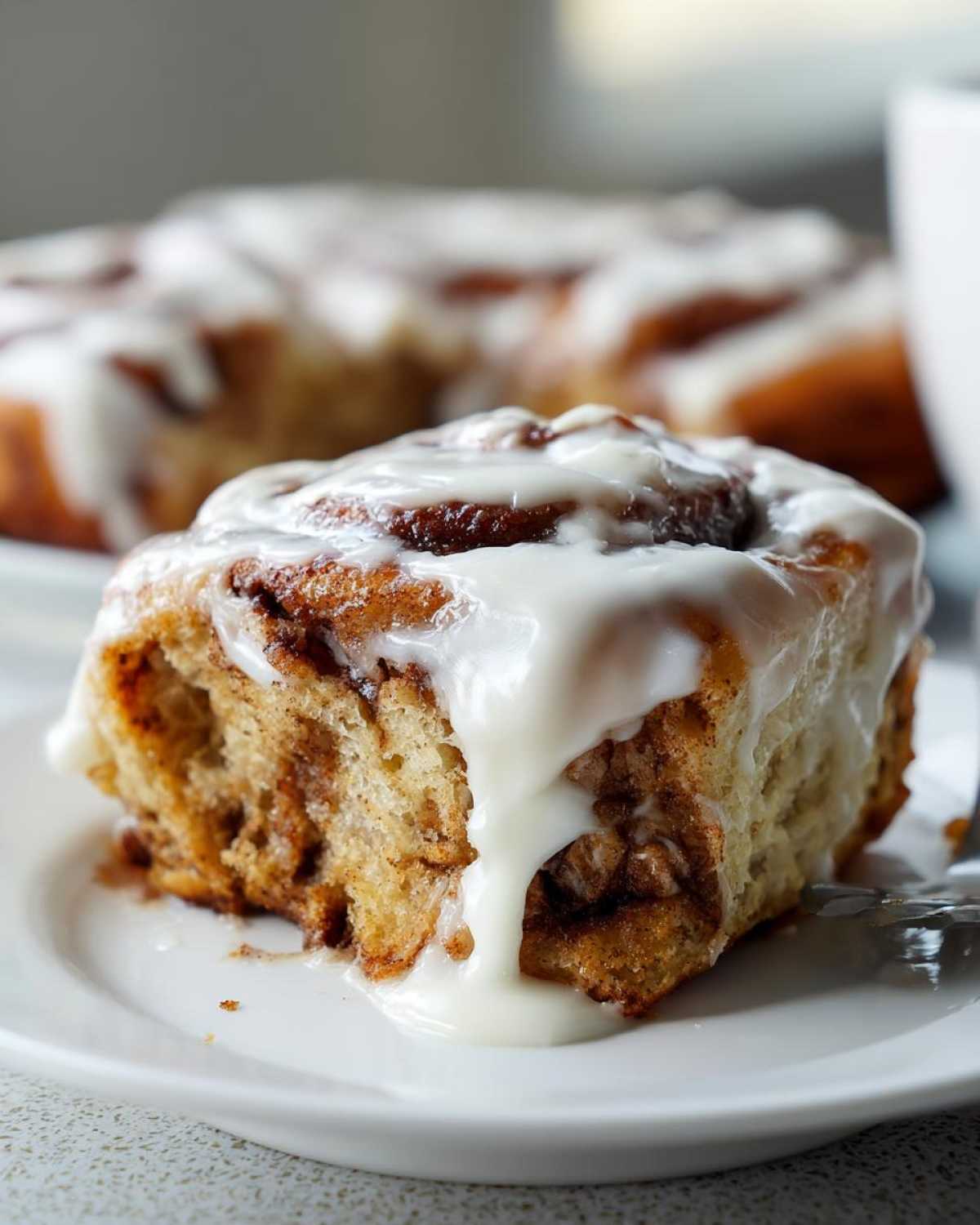 A close-up of a soft, gooey Gluten-Free Cinnamon Roll covered in thick white icing on a white plate.