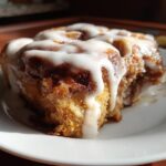 A close-up of a moist, iced Gluten-Free Cinnamon Roll sitting on a white plate, bathed in sunlight.
