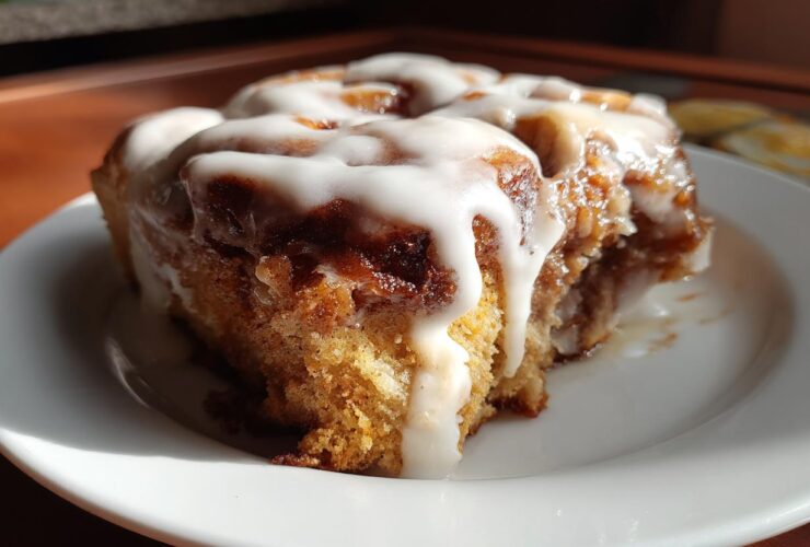 A close-up of a moist, iced Gluten-Free Cinnamon Roll sitting on a white plate, bathed in sunlight.