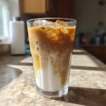 A tall glass of Iced Honey Almond Milk Latte showing layers of white almond milk and rich espresso swirling together over ice.
