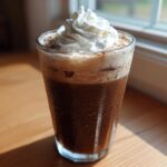 A tall glass of cold Iced Mocha topped with a swirl of whipped cream, sitting on a wooden surface near a window.