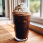 A tall, sweating glass filled with a dark Iced Mocha, topped high with whipped cream.