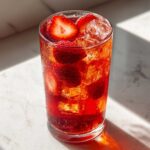 A tall, condensation-covered glass filled with vibrant red Iced Strawberry Lemon Tea, ice cubes, and fresh strawberry slices.