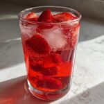 Close-up of a tall glass filled with bright red Iced Strawberry Lemon Tea, ice cubes, and sliced strawberries.