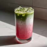 A tall glass showing the beautiful layers of an Iced Strawberry Matcha Latte: bright pink strawberry puree, white milk, and vibrant green matcha.