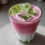 Close-up of a layered Iced Strawberry Matcha Latte with pink foam, ice cubes, and green matcha powder sprinkled on top.
