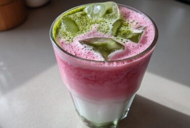 Close-up of a layered Iced Strawberry Matcha Latte with pink foam, ice cubes, and green matcha powder sprinkled on top.