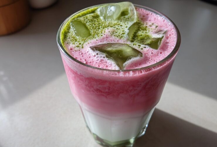 Close-up of a layered Iced Strawberry Matcha Latte with pink foam, ice cubes, and green matcha powder sprinkled on top.