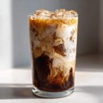 Close-up of a tall glass filled with an Iced Vanilla Chai Latte, showing rich dark chai swirling with white cream.