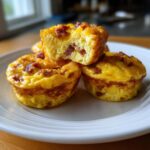 Close-up of three golden Keto Bacon and Egg Muffins on a white plate, one is broken open showing bacon inside.