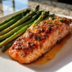 A perfectly cooked fillet of Keto Garlic Butter Salmon glazed with herbs, served alongside bright green asparagus on a white plate.