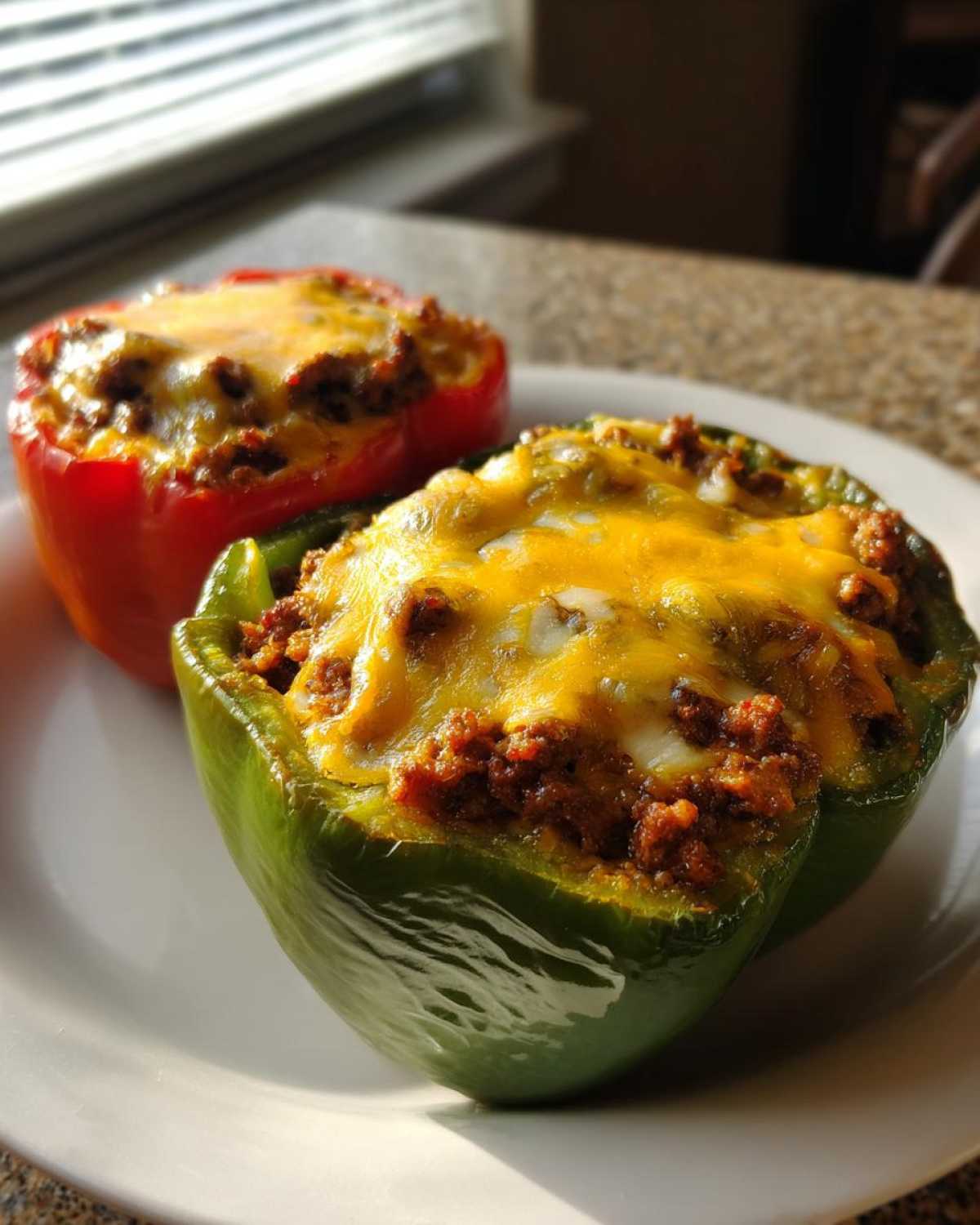 Two Keto Taco Stuffed Peppers, one green and one red, topped with melted cheddar cheese, served on a white plate.